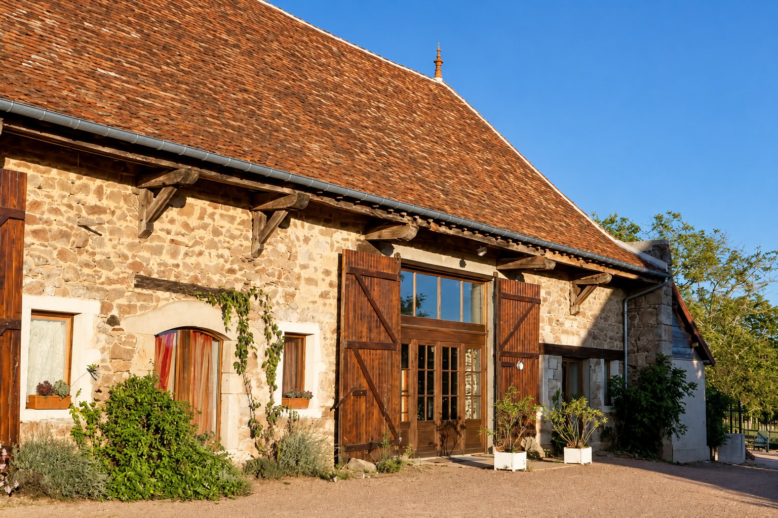 Façade d'une ancienne ferme en pierres apparentes avec un grand toit de tuiles, des volets en bois ouverts et des plantes en pots devant l'entrée, baignée de lumière dorée.