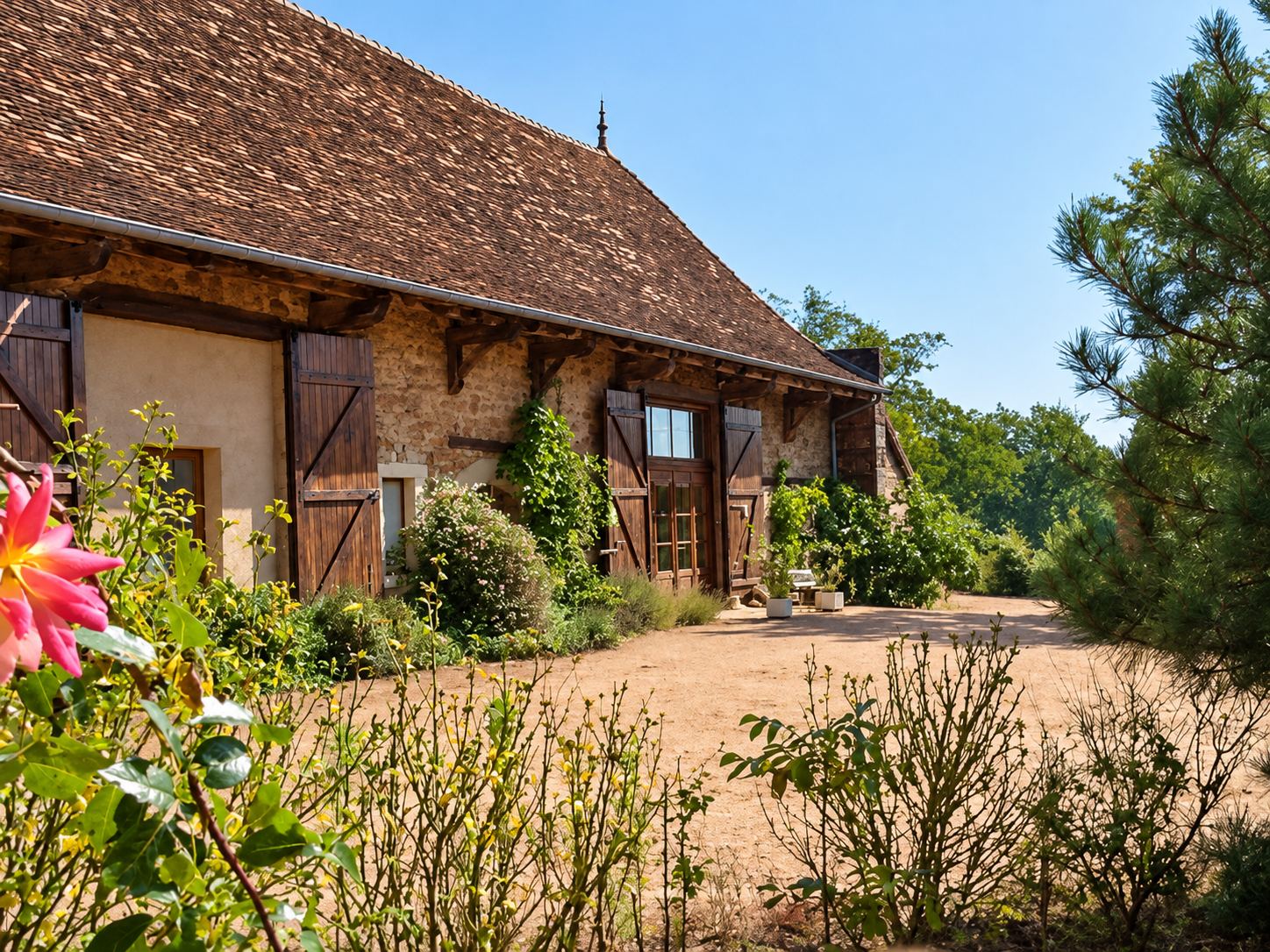 Une longue grange en briques et pans de bois avec un grand toit de tuiles brunes, entourée de massifs fleuris, d'arbres et d'une cour de terre battue ensoleillée.