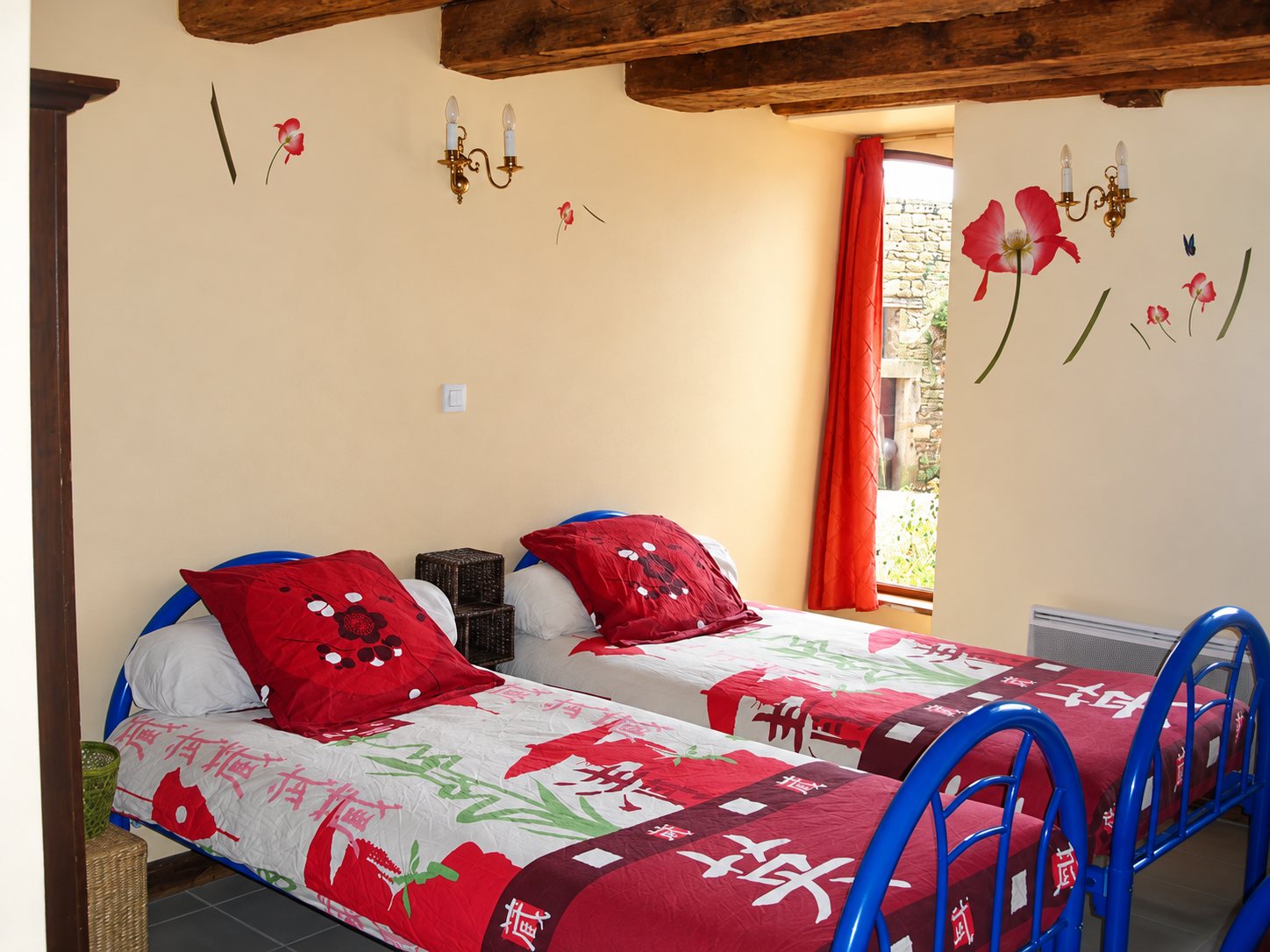 Chambre rustique aux poutres apparentes avec deux lits à armature bleue, couvertures rouges à motifs asiatiques, rideau orange et fleurs décoratives sur les murs.