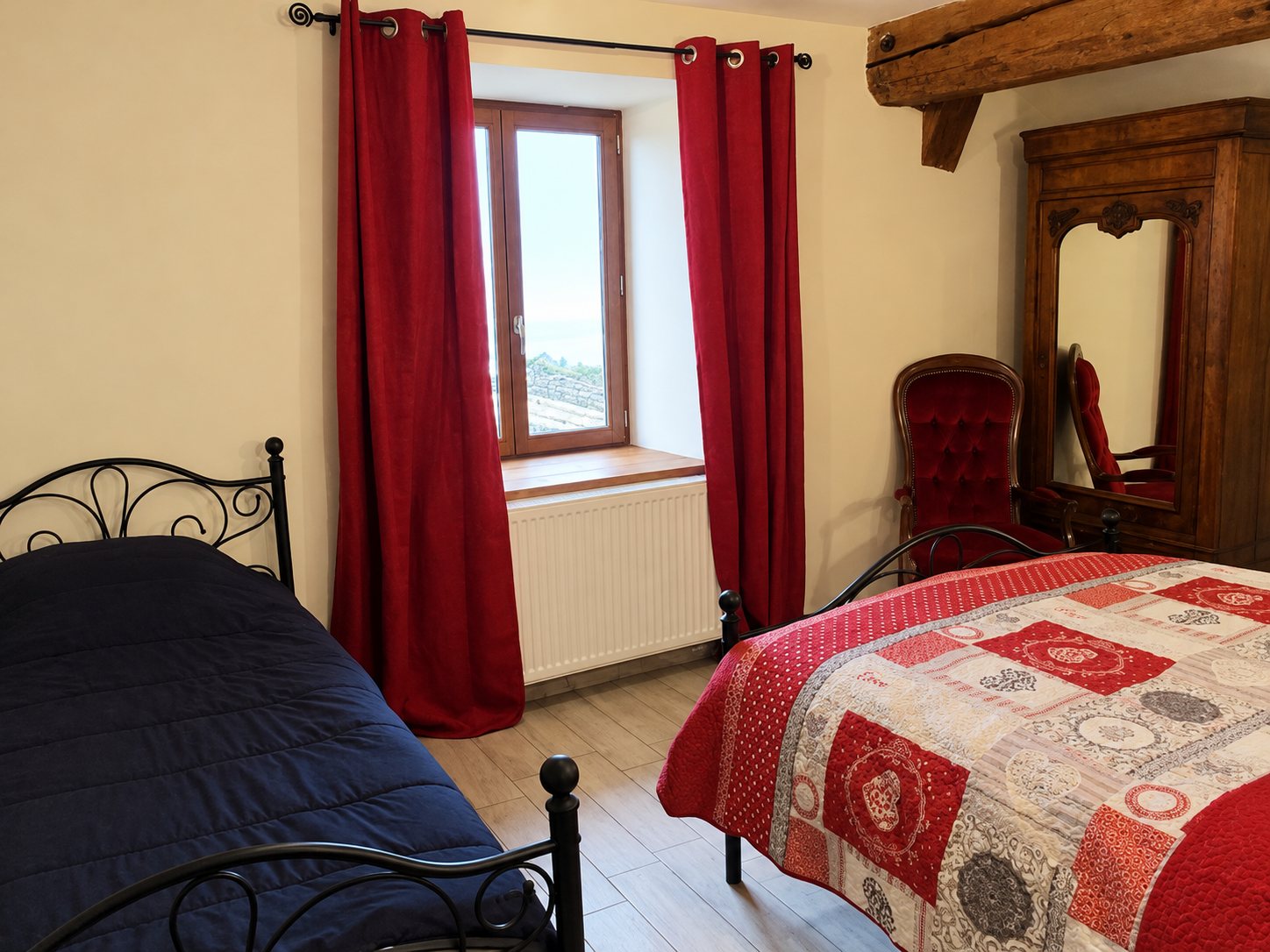 Chambre rustique avec deux lits en fer forgé, un patchwork rouge, des rideaux bordeaux à la fenêtre et une armoire ancienne au fond.