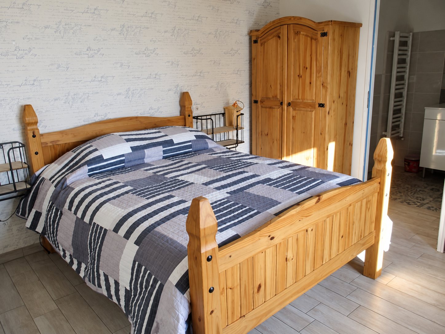 Chambre claire avec un lit double en pin recouvert d'un patchwork bleu et gris, une armoire en bois et une salle de bain en arrière-plan.