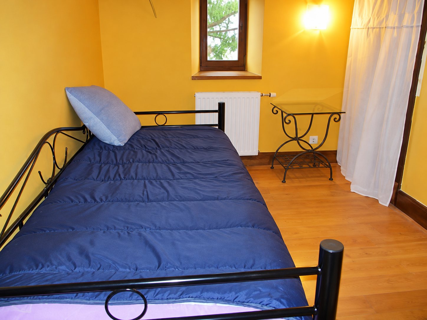 Une chambre aux murs jaunes avec un lit en fer forgé noir, un couvre-lit bleu foncé, une petite table en fer et un parquet en bois.