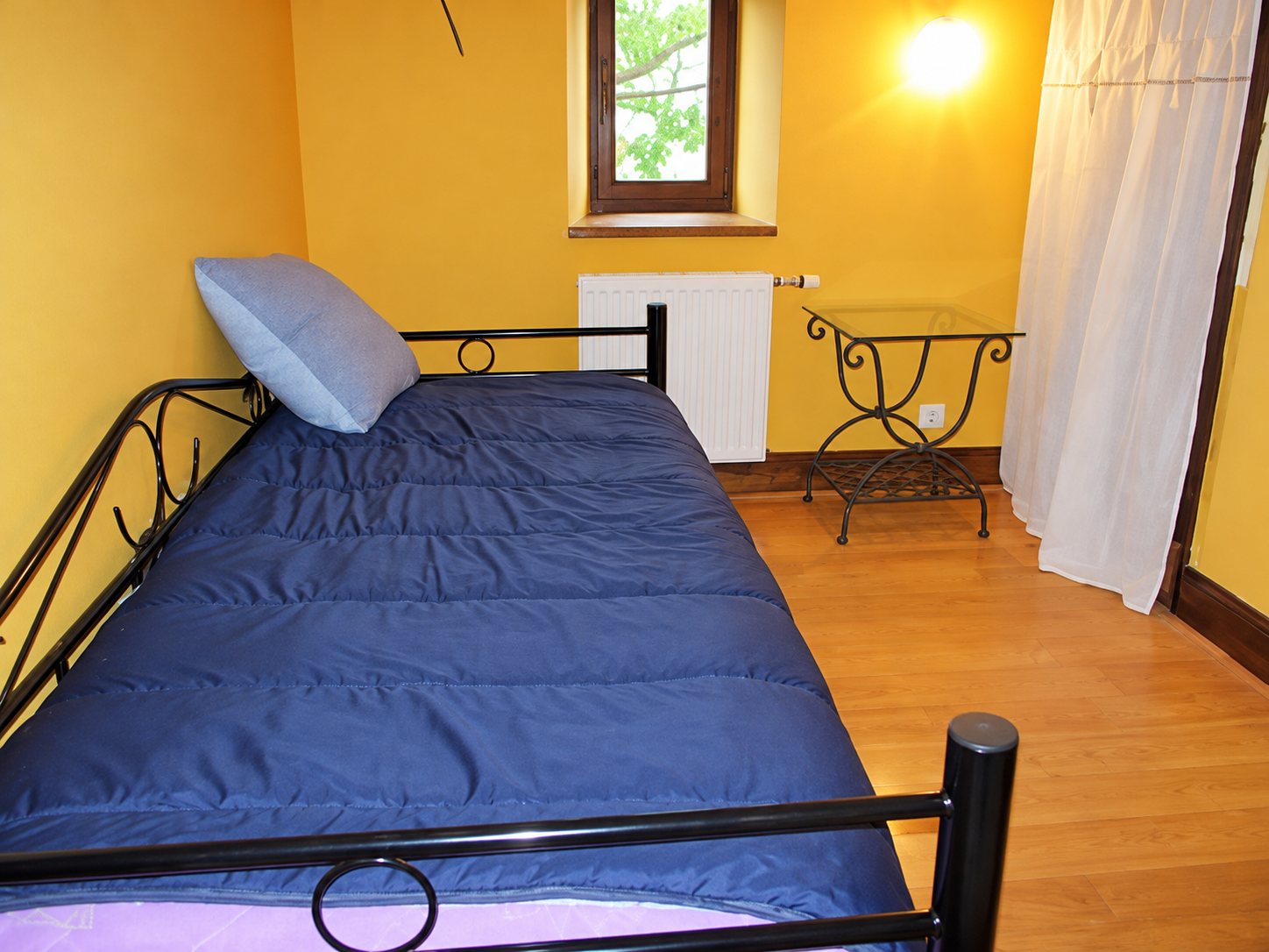 Une chambre aux murs jaunes avec un lit en fer forgé noir recouvert d'un couvre-lit bleu, une petite table en fer forgé, une fenêtre et un parquet en bois.