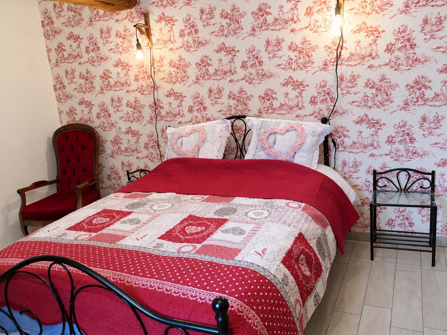 Chambre avec un lit en fer forgé couvert d'un patchwork rouge et blanc à motifs de cœurs, devant un mur tapissé de toile de Jouy rouge, avec un fauteuil rouge et une table de chevet métallique.