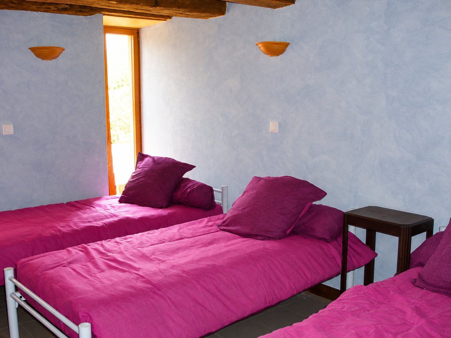 Chambre rustique avec plusieurs lits simples aux draps et coussins violets, murs bleu-gris patinés, poutres apparentes et fenêtre très lumineuse.