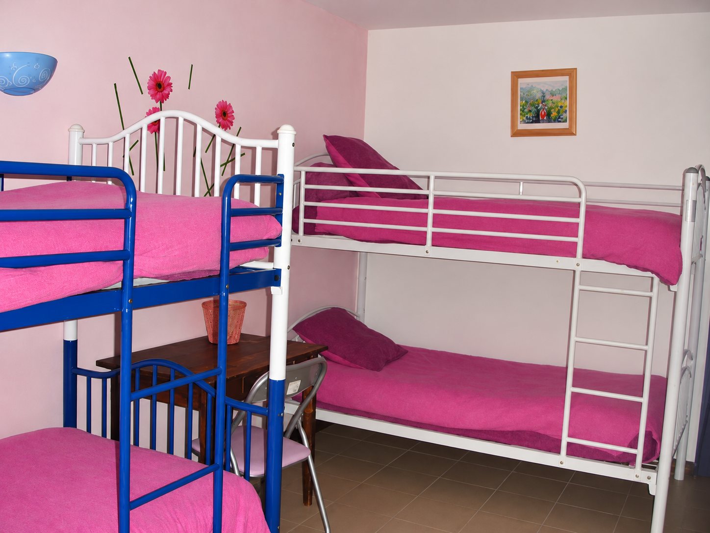 Chambre aux murs clairs avec deux lits superposés, l'un bleu et l'autre blanc, dotés de draps roses, une petite table en bois et un tableau au mur.