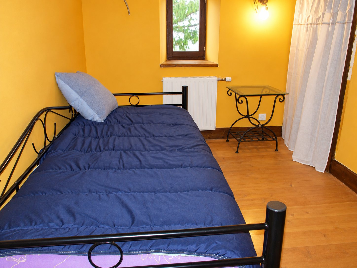 Chambre aux murs jaunes avec un lit en métal noir recouvert d'une couette bleue marine, une table de chevet en verre et fer forgé, un radiateur et une fenêtre.