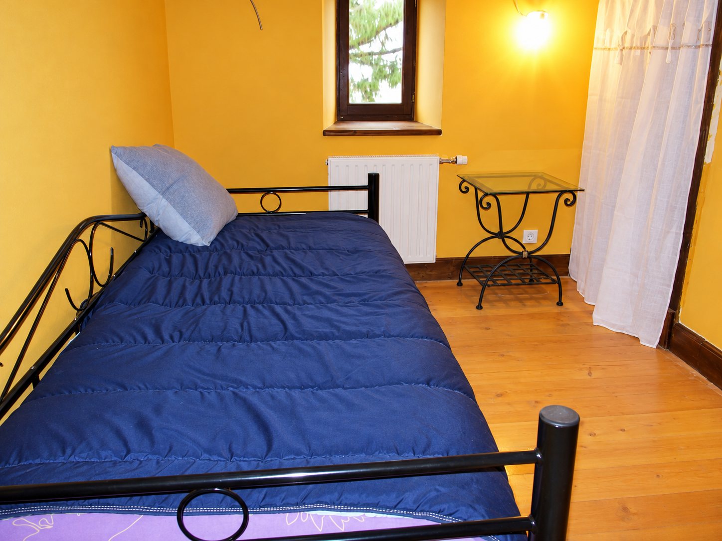 Une chambre aux murs orange avec un lit en métal noir recouvert d'une couette bleu foncé, une table de chevet en verre et fer forgé, un radiateur et un rideau blanc près de la fenêtre.
