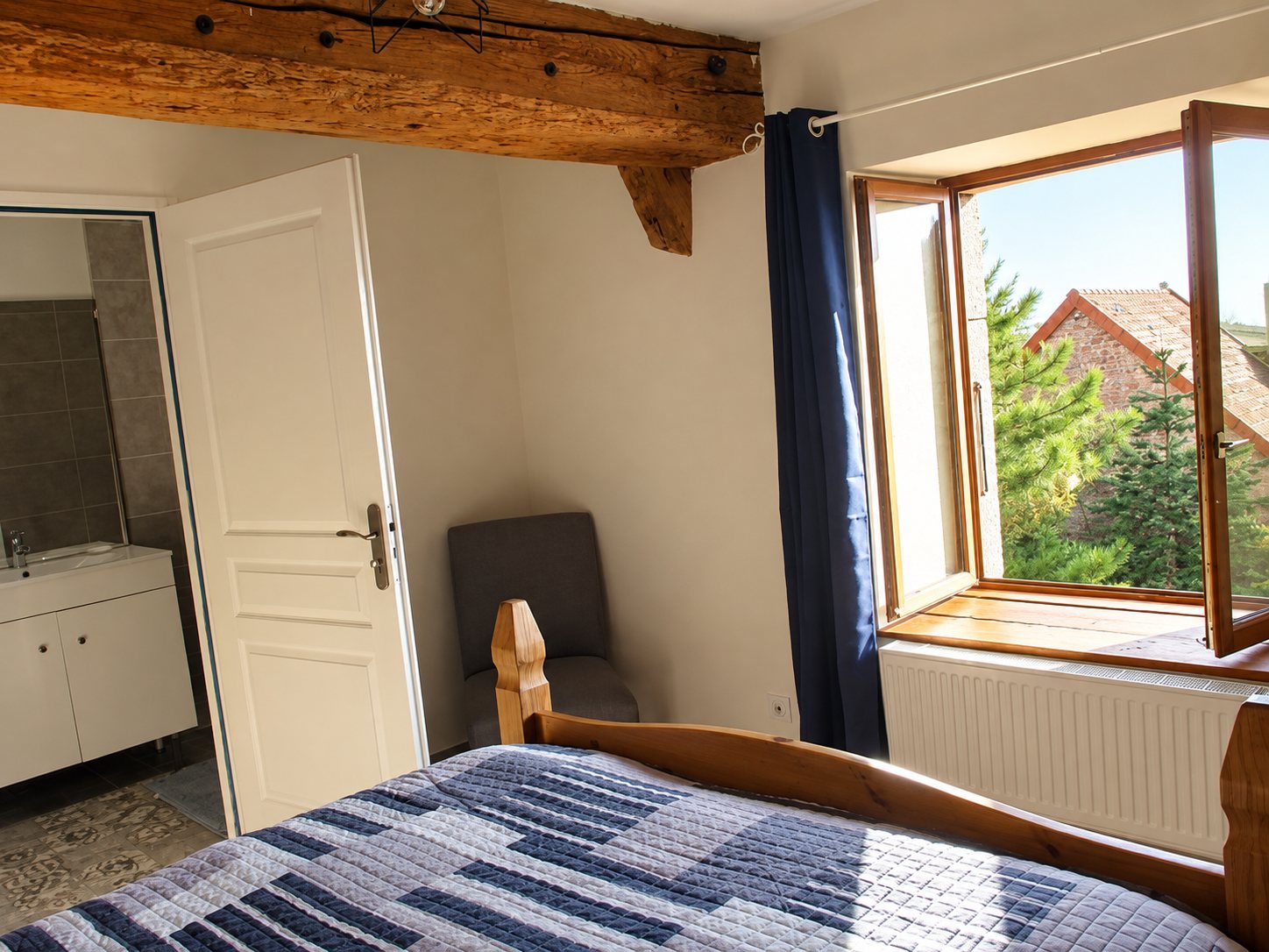 Chambre lumineuse avec une poutre en bois, un lit à courtepointe bleue, une fenêtre ouverte sur des sapins et une salle d'eau attenante.