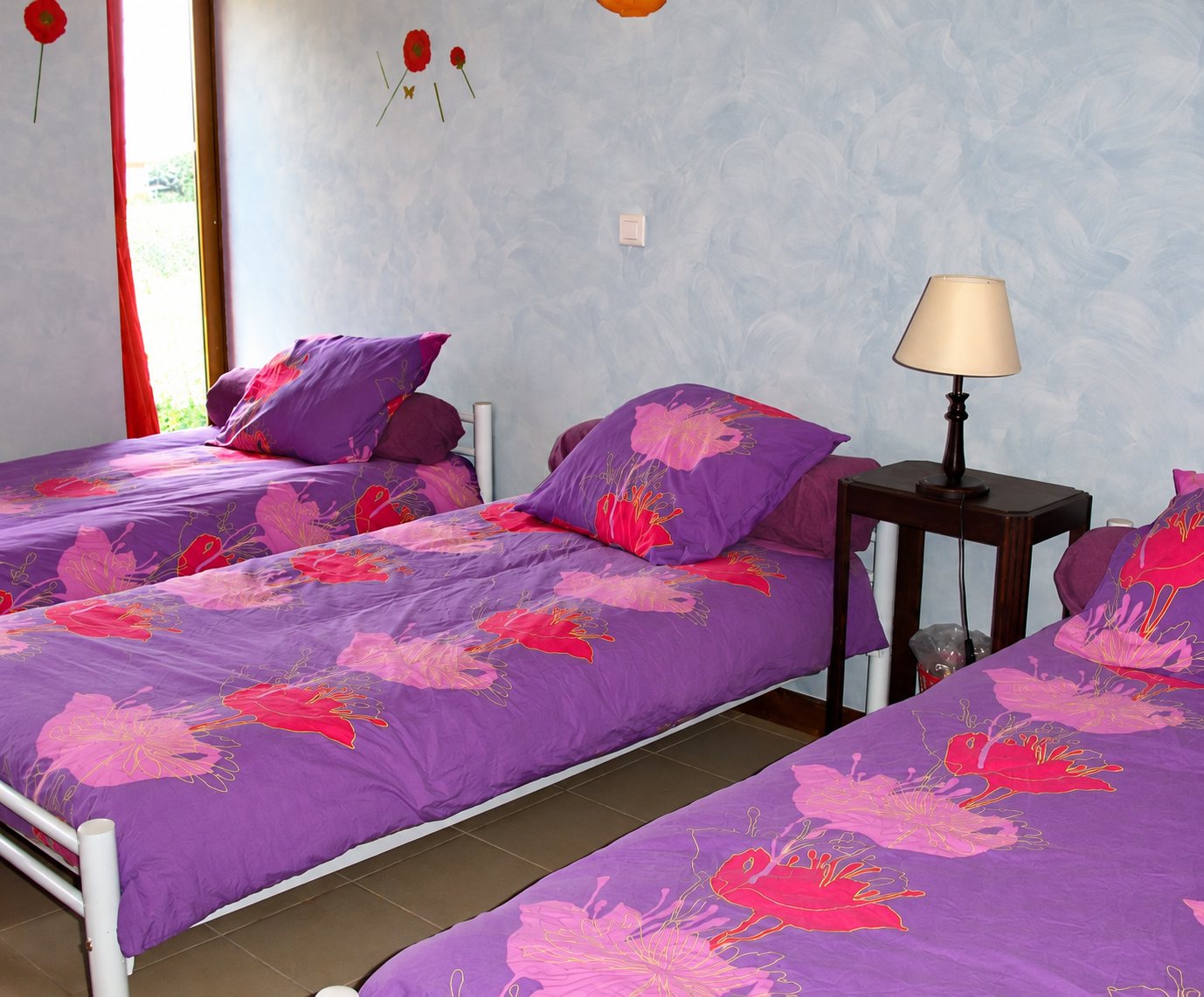 Chambre aux murs patinés bleu-gris avec trois lits simples dotés de housses de couette violettes à motifs de fleurs roses et rouges, et une table de chevet avec lampe.