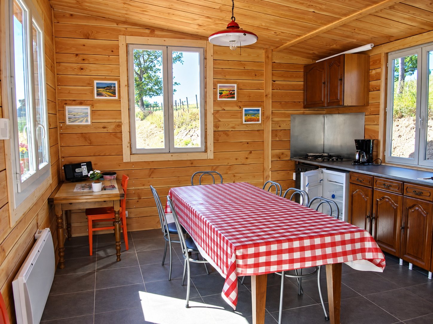 Intérieur lumineux d'un chalet en bois avec une cuisine équipée, une grande table couverte d'une nappe à carreaux rouges et blancs et des fenêtres donnant sur la nature.