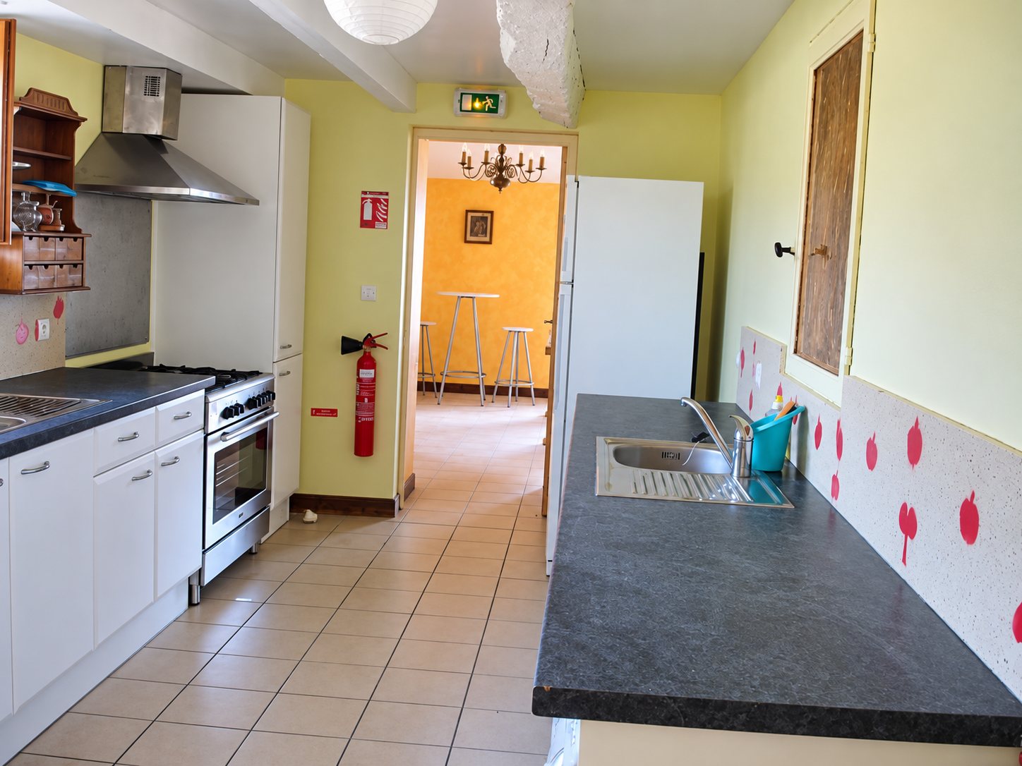 Une cuisine aux murs jaunes avec meubles blancs, cuisinière, évier et extincteur, ouverte sur une salle orange avec tables hautes et tabourets.