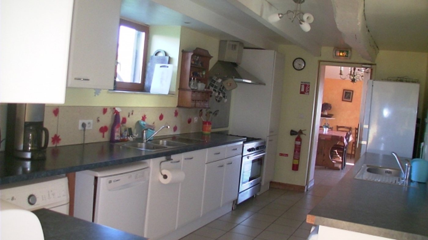 Cuisine lumineuse aux meubles blancs et plans de travail sombres, avec cuisinière, lave-vaisselle et extincteur, ouverte sur une salle à manger orange.