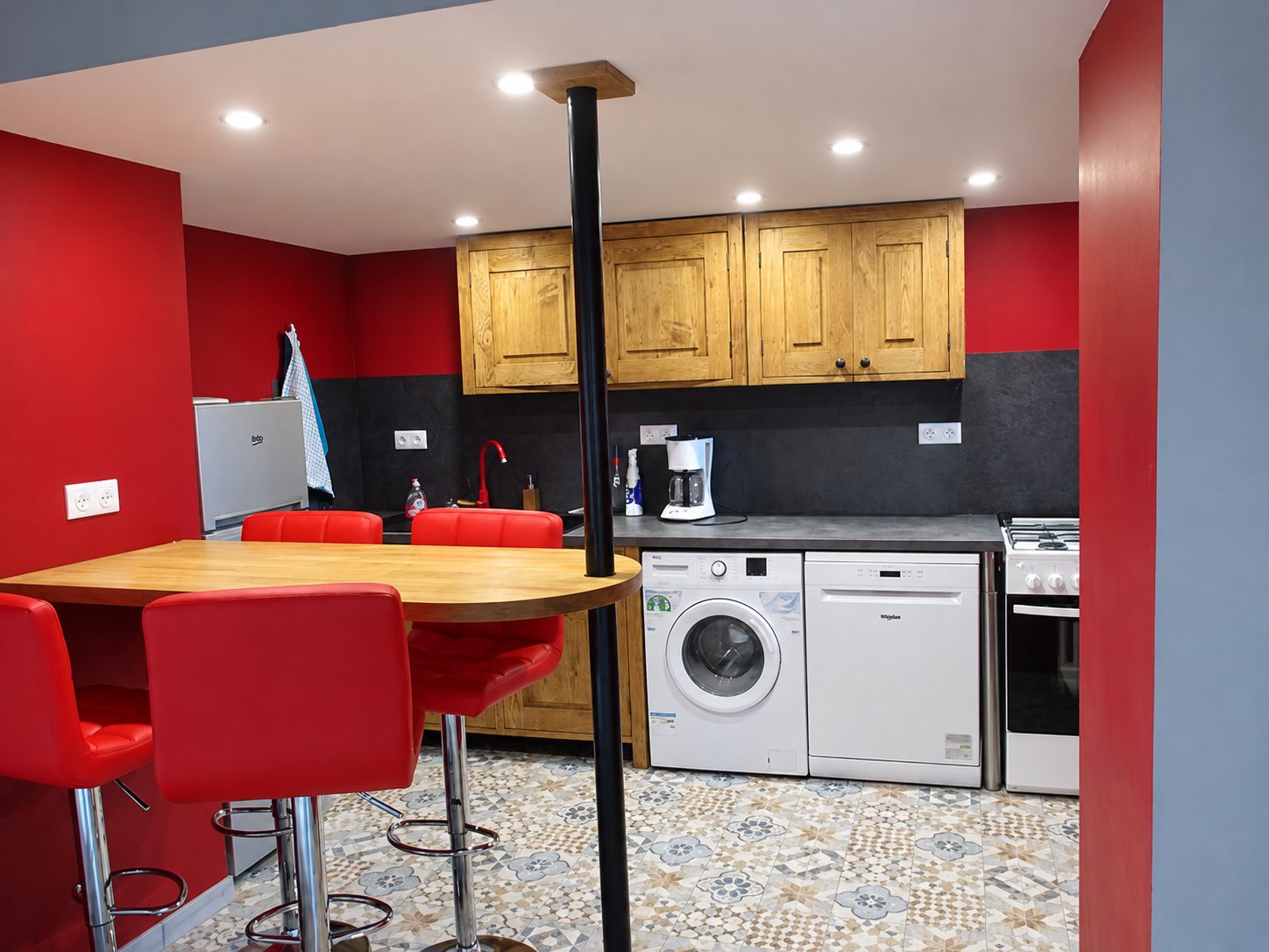 Cuisine équipée aux murs rouges, avec placards en bois, lave-linge, lave-vaisselle, cuisinière et table haute entourée de tabourets rouges sur un carrelage à motifs.