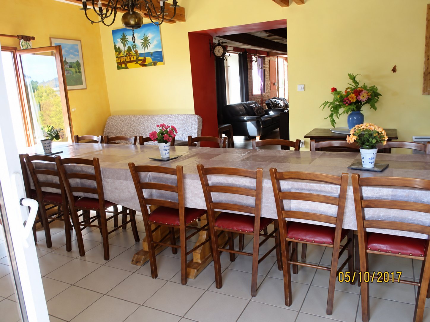 Une grande salle à manger aux murs jaunes avec une longue table entourée de chaises en bois à assise rouge, décorée de bouquets de fleurs.