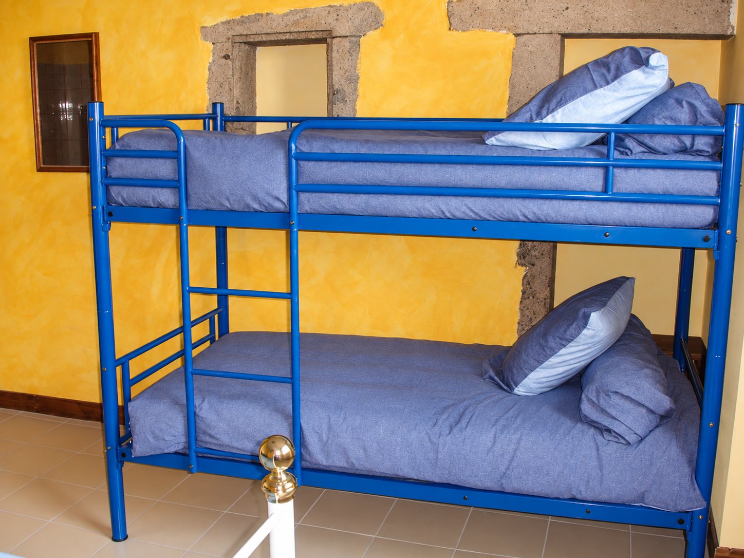 Un lit superposé en métal bleu avec literie grise est installé dans une chambre aux murs jaunes patinés et aux encadrements de pierre.