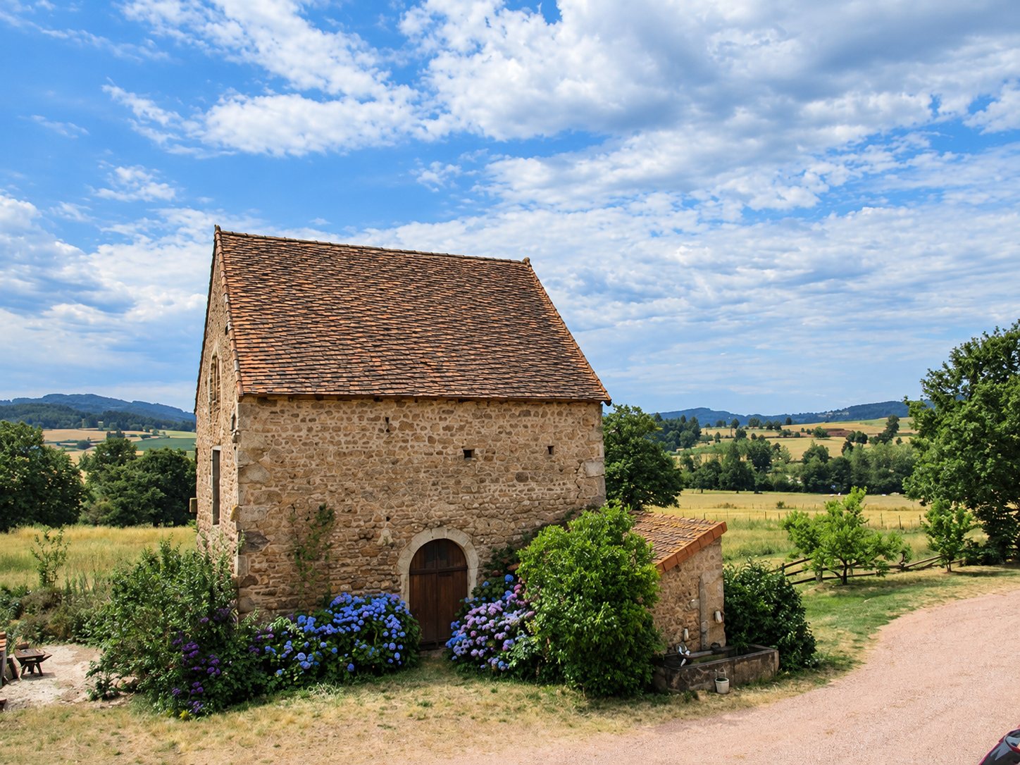 Une ancienne bâtisse en pierre au toit de tuiles, entourée d'hortensias bleus, au milieu d'une campagne vallonnée sous un ciel nuageux.