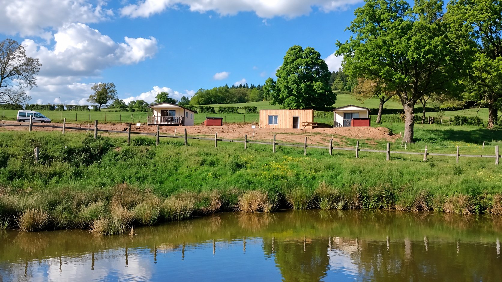 Trois petits chalets en bois et crépi se trouvent sur un terrain de terre rouge, derrière une clôture et un étang, au milieu de prairies vertes.
