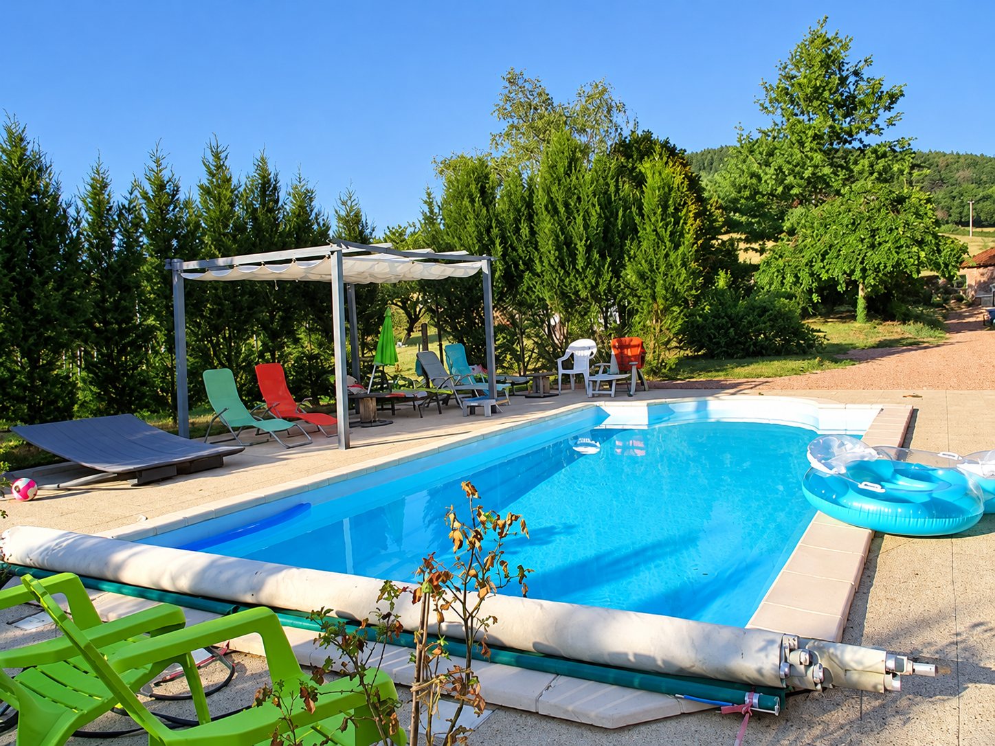 Une piscine rectangulaire à l'eau bleue avec sa bâche enroulée, entourée de transats colorés, d'une pergola et d'arbres sous un ciel dégagé.