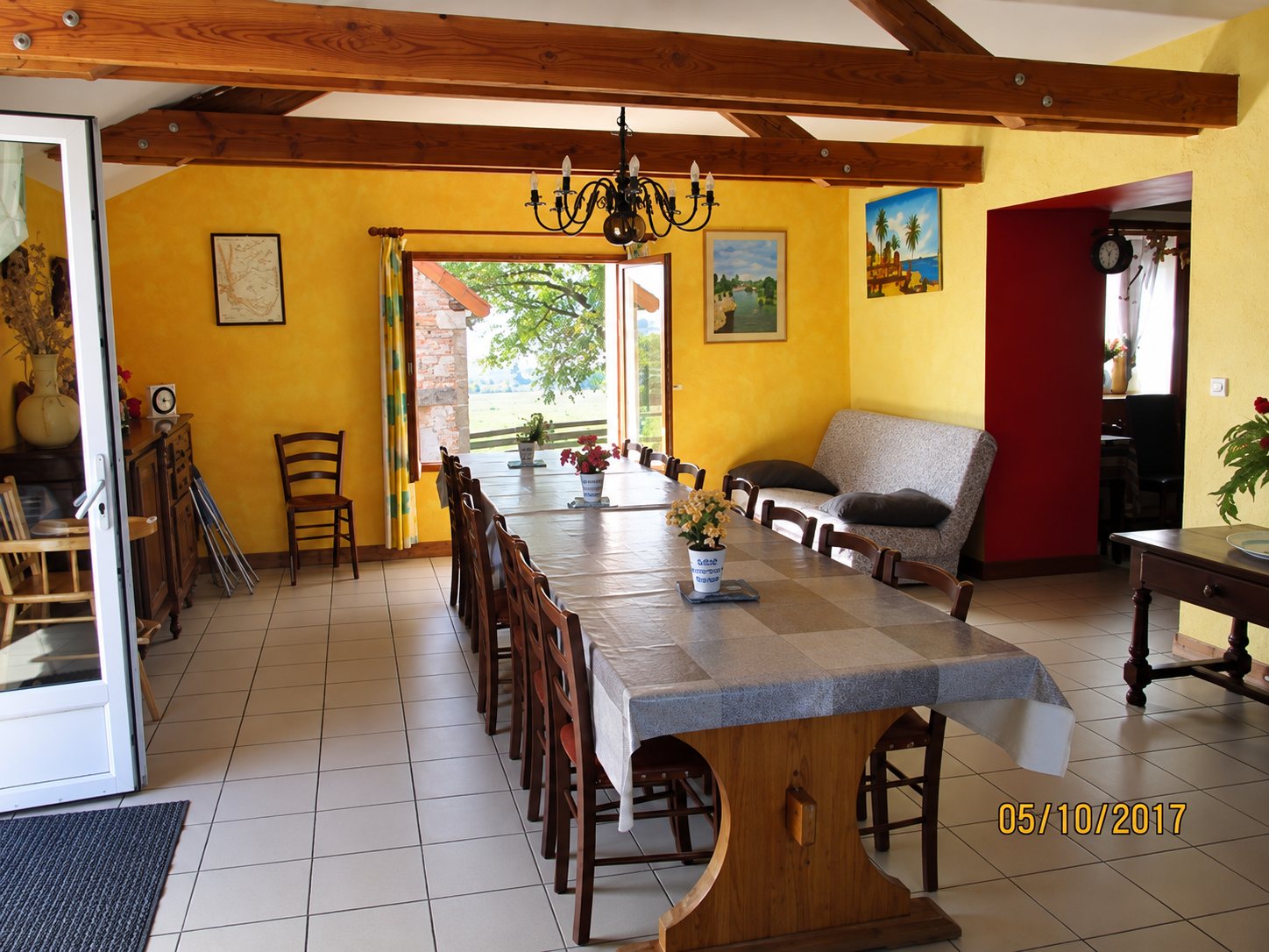 Grande salle à manger aux murs jaunes avec poutres en bois, une longue table recouverte d'une nappe grise entourée de chaises et un lustre au plafond.