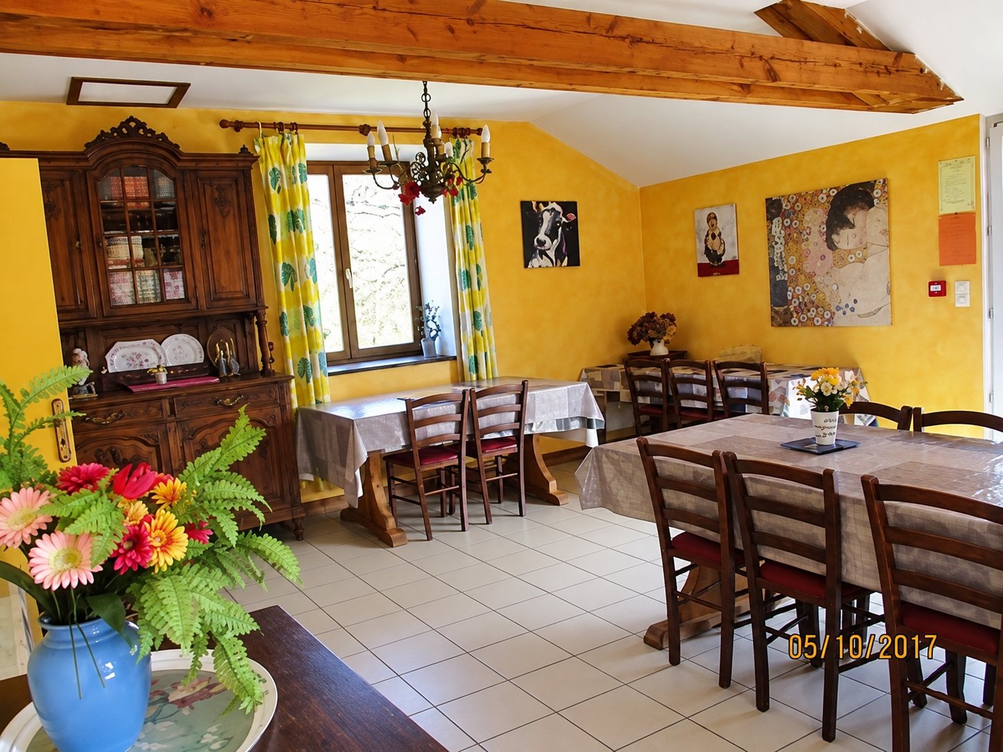 Salle à manger lumineuse aux murs jaunes avec poutres apparentes, grandes tables en bois, chaises, vaisselier ancien et bouquet de fleurs au premier plan.