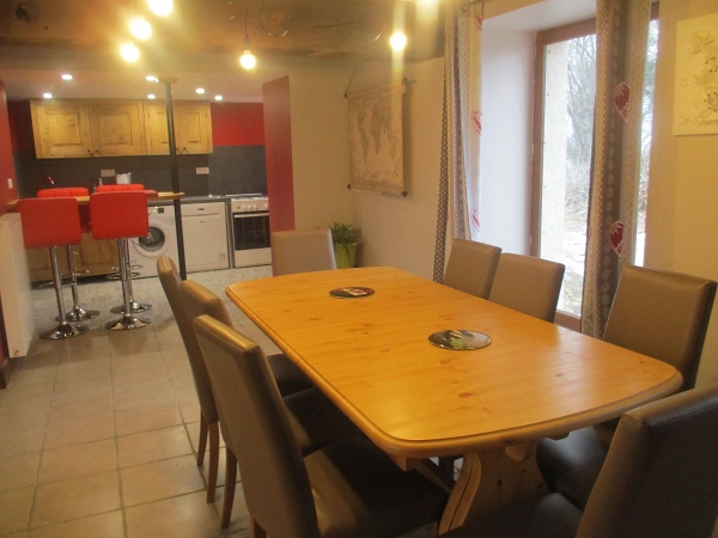 Une grande table en bois clair entourée de chaises beiges dans une pièce ouverte sur une cuisine aux murs rouges avec tabourets rouges.