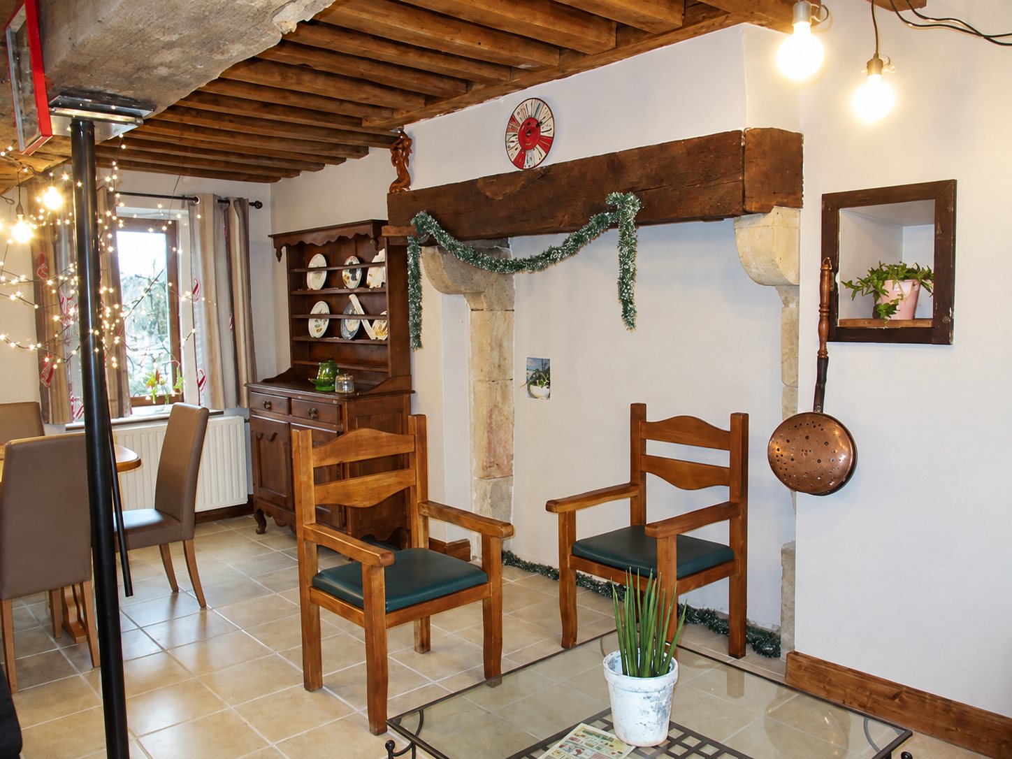 Intérieur rustique avec deux fauteuils en bois, une table basse en verre, un vaisselier ancien et une cheminée en pierre décorée d'une guirlande.
