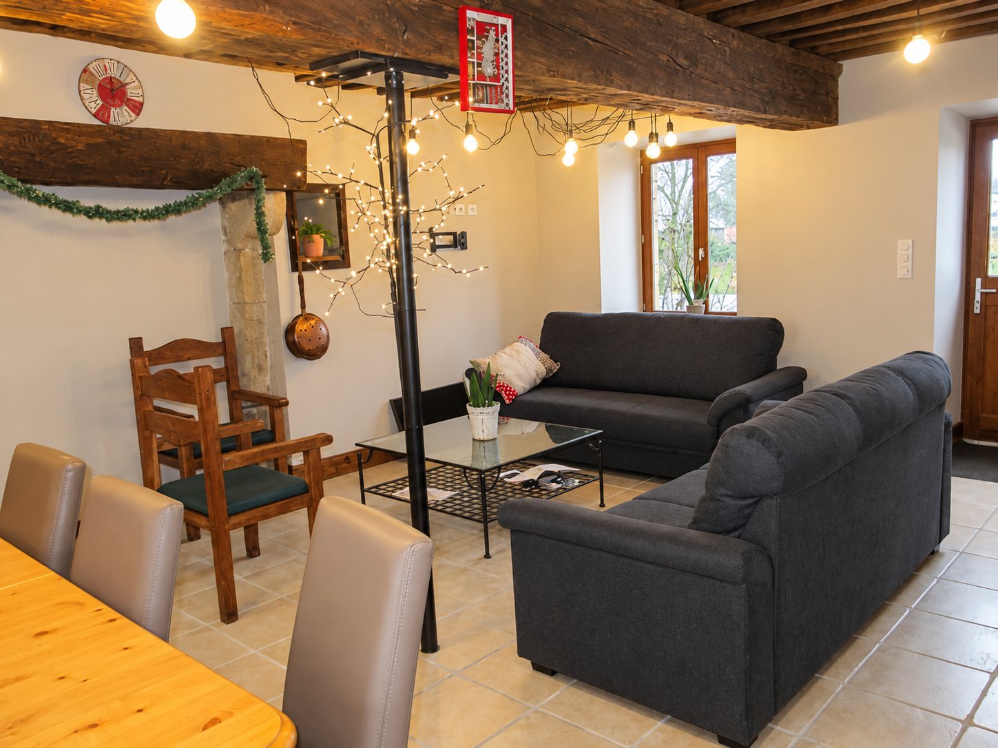 Salon rustique avec deux canapés gris, une table basse en verre, une chaise en bois et une table à manger, sous des poutres apparentes décorées de guirlandes lumineuses.