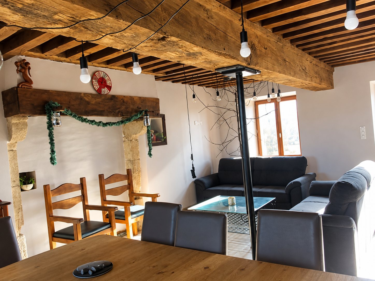 Pièce de vie rustique avec grosses poutres en bois, cheminée en pierre ornée d'une guirlande, chaises en bois, canapés gris et table basse en verre.