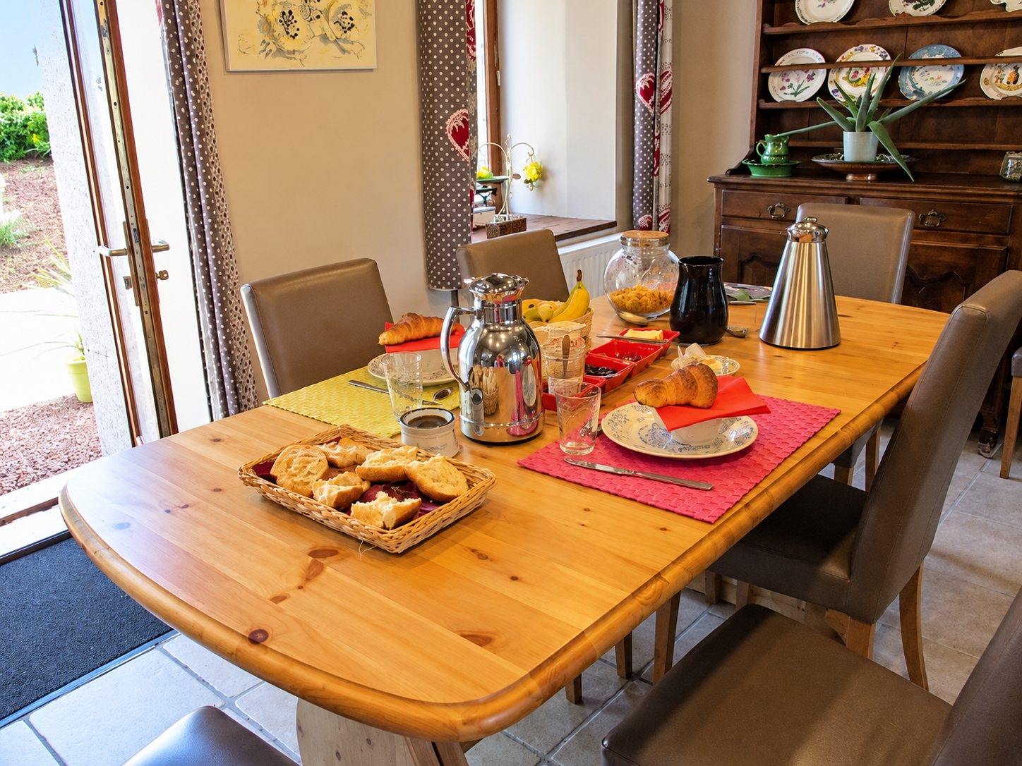 Une table en bois dressée pour le petit-déjeuner avec pain, viennoiseries, confiture et thermos, près d'un vieux buffet garni d'assiettes décoratives.