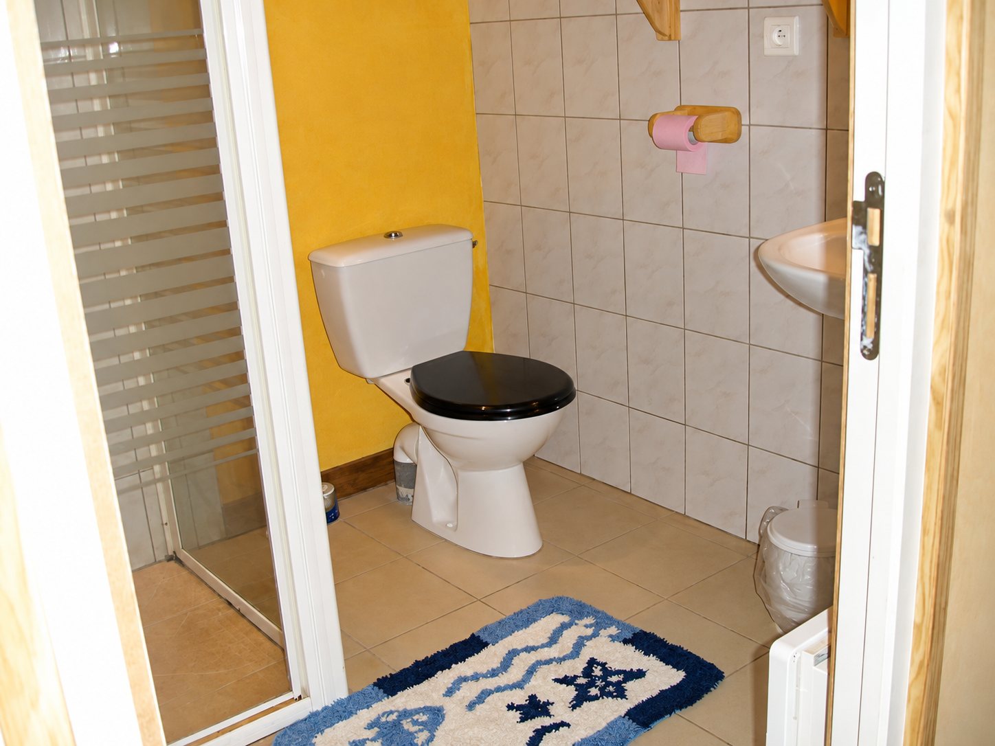 Des toilettes avec un siège noir, un mur jaune, un lavabo, un carrelage beige et un tapis bleu et blanc au sol, à côté d'une douche vitrée.